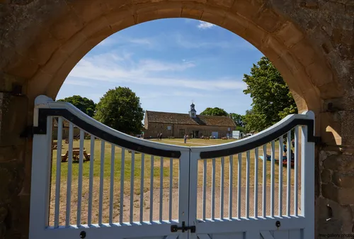This architectural landscape photograph shows the yard of Hardwick Hall, a prominent Elizabethan manor house in Derbyshire, United Kingdom. Taken on a summer morning, the image is framed through a stone archway and light blue gate that opens onto a grassy yard with picnic tables and people enjoying the outdoors. In the background, the distinctive clock tower and stone-built stables associated with the National Trust site are visible, set amongst mature trees under a blue sky. Hardwick Hall, renowned for its Elizabethan architecture, is a notable historical landmark managed by the National Trust, reflecting the heritage of Derbyshire and the United Kingdom.